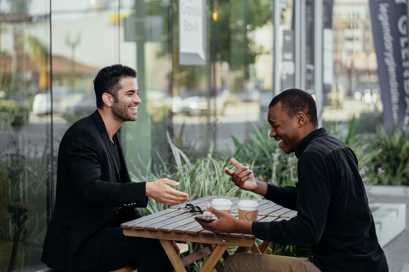 Deux personnes qui rient en pleine conversation animée, gestuelle vive, autour d'une table de café en terrasse