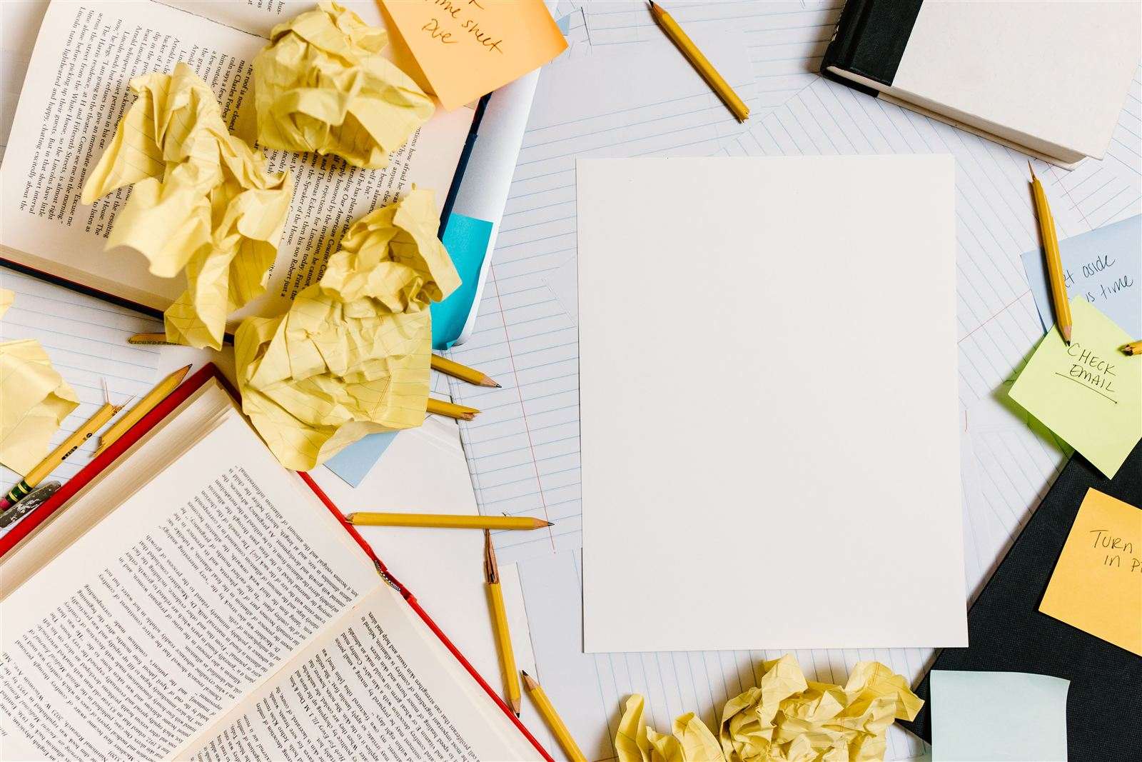 Un bureau encombré de boules de papier froissé, de notes autocollantes et de livres ouverts, avec une page blanche au centre, symbole d'un roman dont l'organisation s'est éparpillée